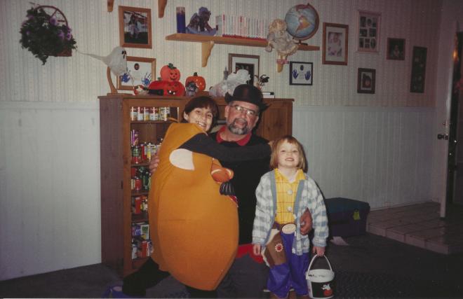 Family Halloween photo from the 90s. Sister dressed as a pumpkin, dad in a dark costume with a cowboy hat, and a small Shannon dressed as Woody from Toy Story holding a pumpkin candy bucket.